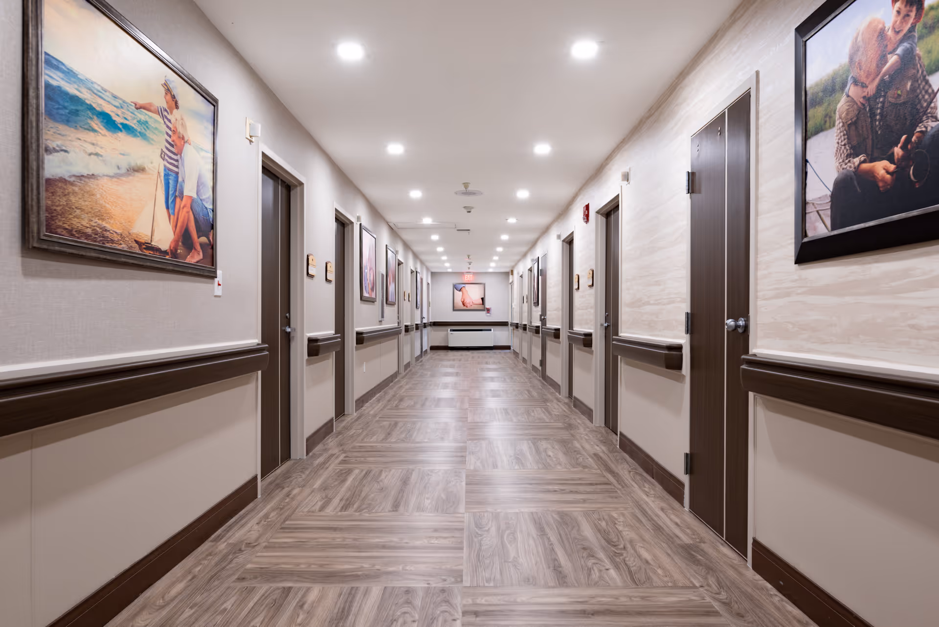 A well-lit hallway in a senior living facility with wood-patterned flooring and beige walls. The hallway has multiple closed doors on both sides, handrails along the walls, and framed pictures hanging above the handrails. The ceiling has recessed lighting evenly spaced along the corridor.