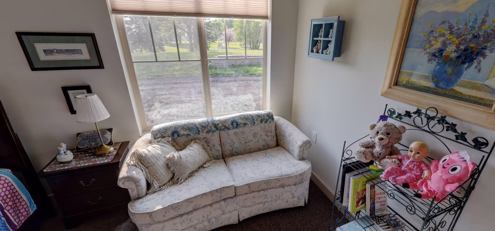 A small sitting area with a floral loveseat beneath a window, a side table with a lamp, and a wire shelf holding stuffed animals and books.