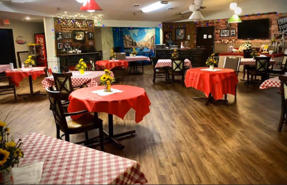 Dining room with round tables draped in red and red-and-white checkered tablecloths, sunflower centerpieces, and wood flooring.