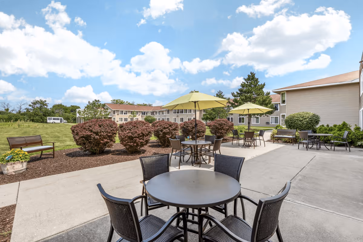 Outdoor patio area at Layton Terrace Senior Living with round tables and chairs, some shaded by green umbrellas. The patio is surrounded by bushes and greenery, with a clear blue sky and a multi-story building in the background.