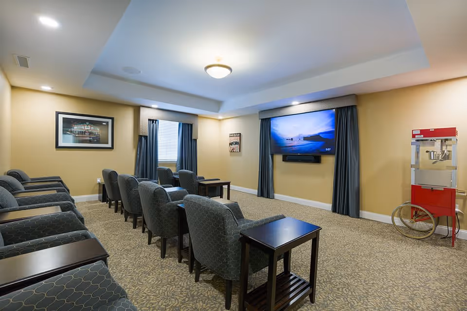 A cozy media room with two rows of cushioned chairs facing a large flat-screen TV mounted on a beige wall. The room has carpeted flooring, a popcorn machine on the right side, framed pictures on the walls, and windows with blue curtains.