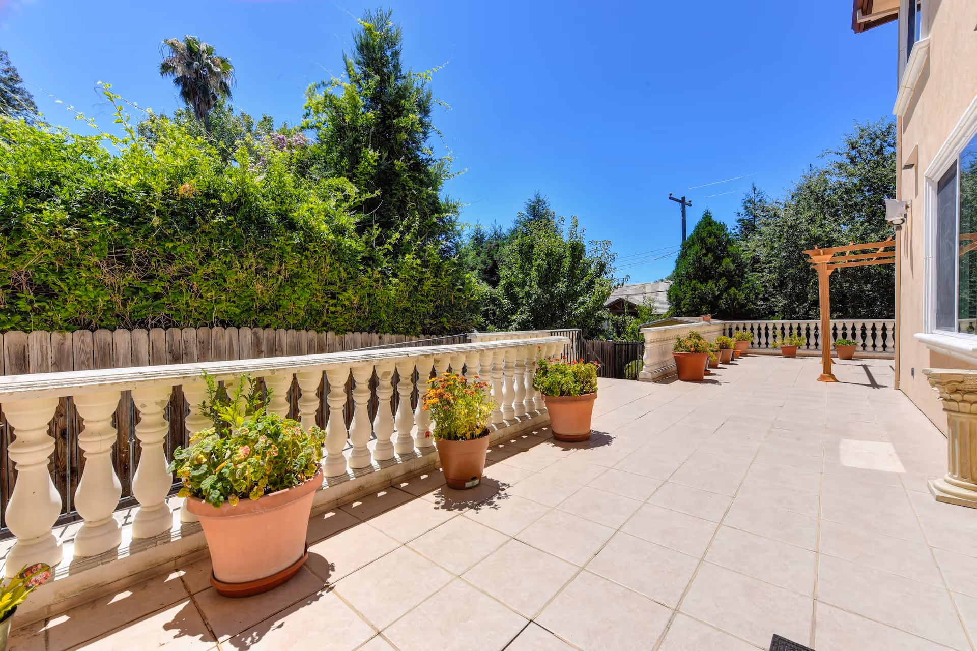A spacious outdoor patio area with beige tiled flooring, white balustrade railing, several potted plants along the edge, and a clear blue sky overhead. There are trees and greenery surrounding the patio, and part of a building with a window and a pergola is visible on the right side.
