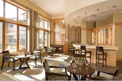 A bright and spacious common area with large windows letting in natural light. The room features several small round tables with chairs arranged around them, and a bar counter with high stools. The decor includes light wood cabinetry and neutral tones, creating a warm and inviting atmosphere.