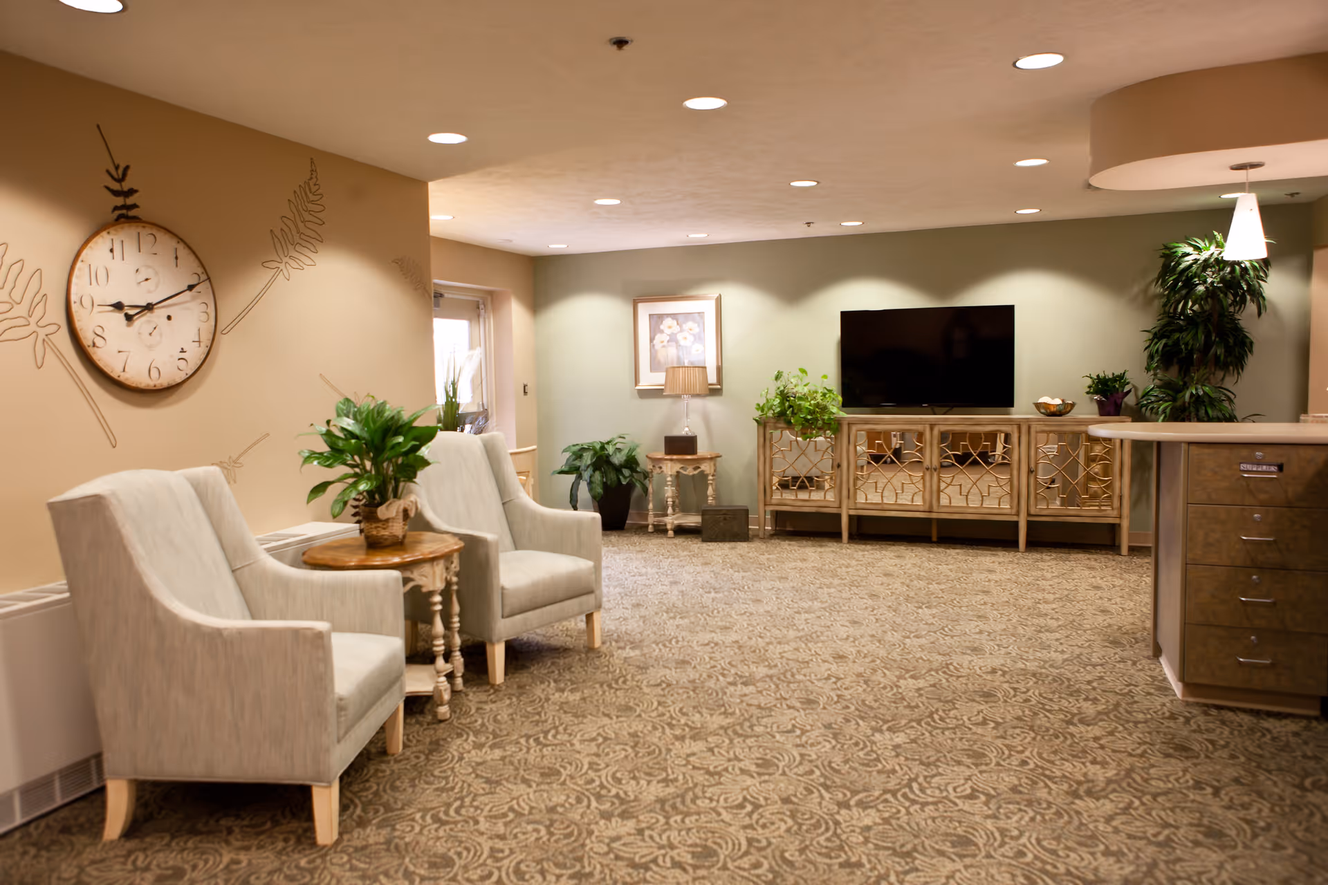 A cozy living room area in a senior living facility featuring two upholstered armchairs with a small wooden table and a potted plant between them. A large wall clock and decorative wall art are on the left wall. The room has a patterned carpet, a flat-screen TV mounted on a green accent wall, a mirrored cabinet below the TV, and various plants and a table lamp adding to the decor.