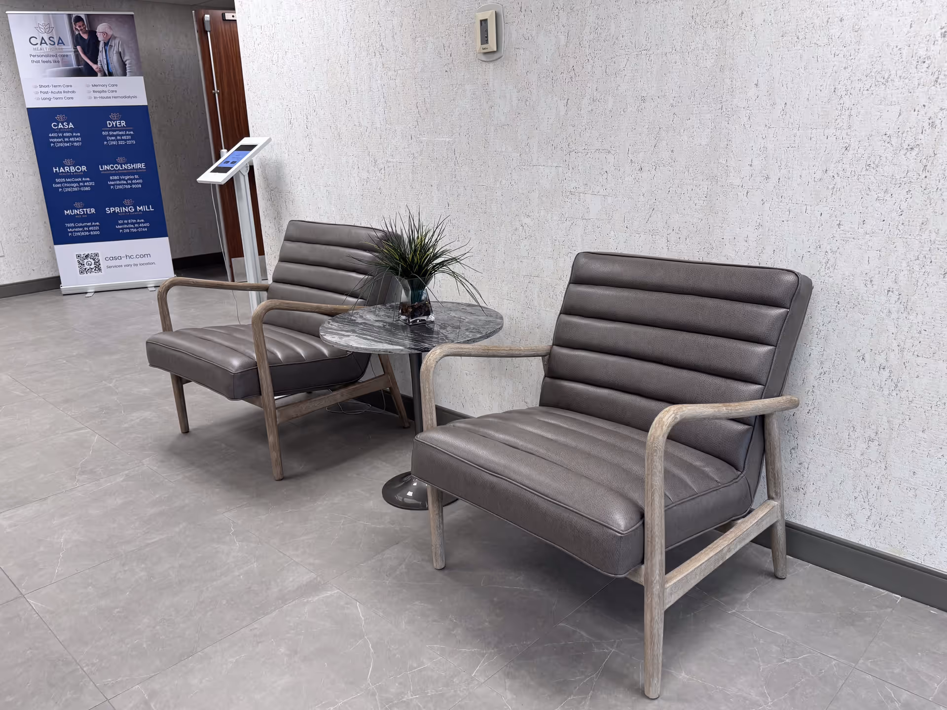Two gray upholstered chairs flanking a small round table with a potted plant in a facility waiting area.