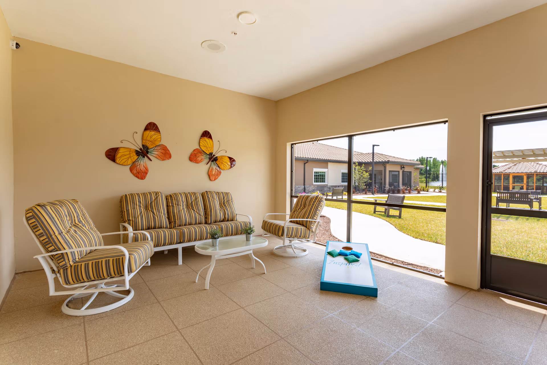 Covered patio area with striped cushioned chairs and a sofa around a small white coffee table. Two large decorative butterfly wall hangings are on the beige wall. Outside, there is a grassy area with benches and a building with a tiled roof visible through large open windows and a door.