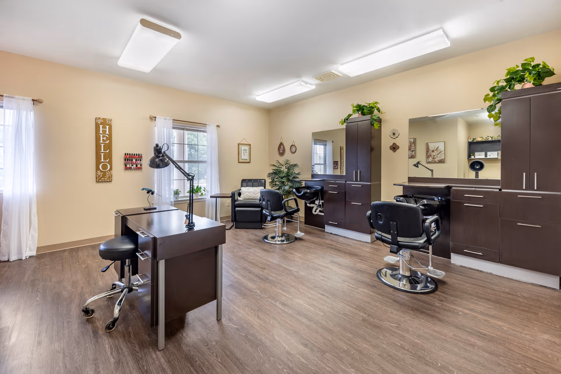 A bright and clean salon room with two black salon chairs in front of large mirrors and dark wood cabinetry. There is a manicure table with a black lamp and stool in the foreground. The room has light beige walls, wood flooring, and two windows with sheer white curtains letting in natural light. Decorative plants and wall art add a welcoming touch.