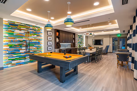 Bright communal activity room with a pool table, dining table, kitchenette, colorful wall art, and pendant lighting.
