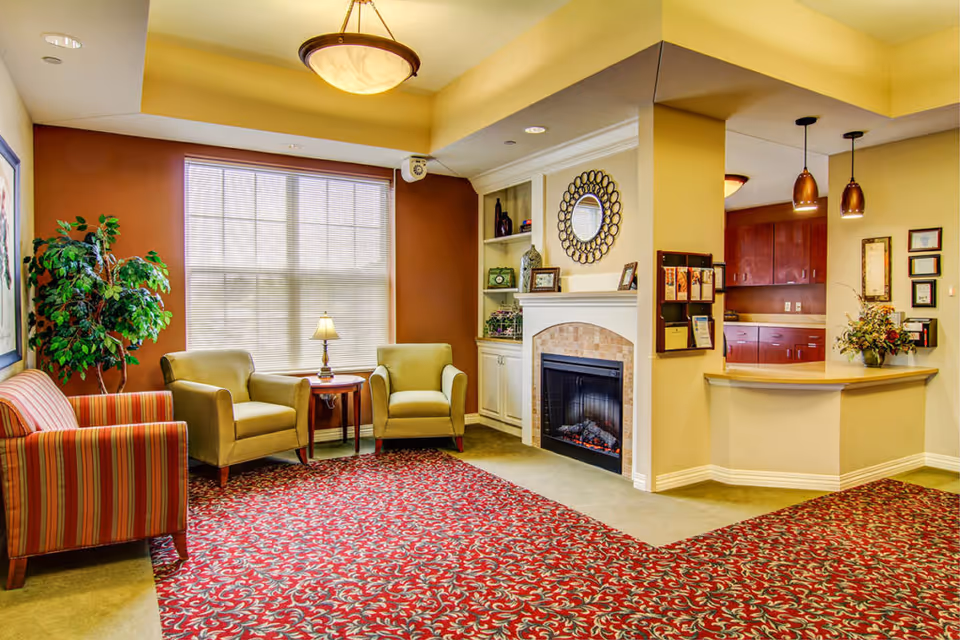 Cozy senior living facility sitting area with armchairs, a fireplace, a reception desk, and patterned red carpet.