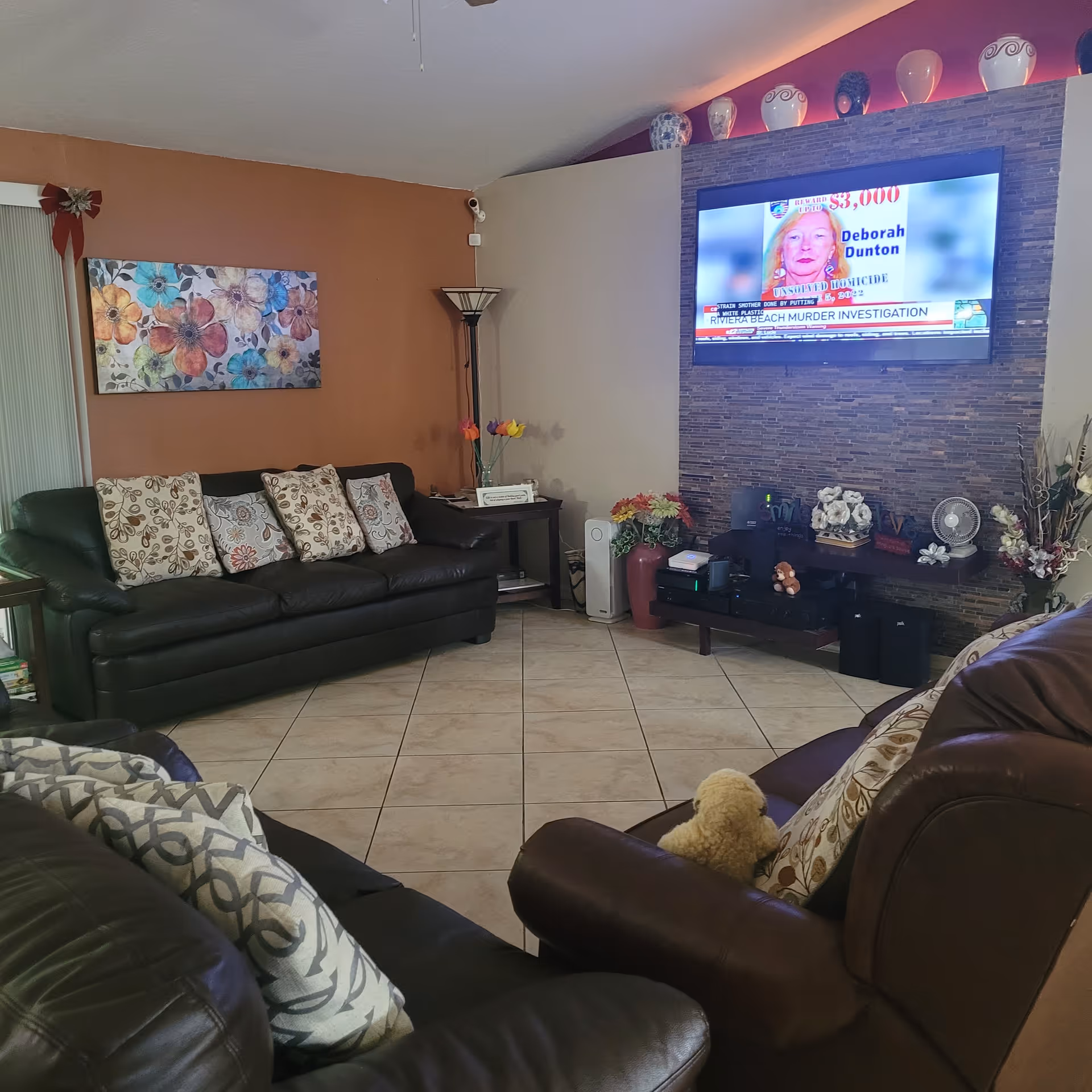 A cozy living room with brown leather sofas adorned with patterned cushions. A flat-screen TV is mounted on a stone accent wall, displaying a news segment. The room has tiled flooring, a side table with a vase of flowers, and decorative items on a low shelf beneath the TV. The walls are painted in warm tones, and there is a colorful floral painting on one wall.