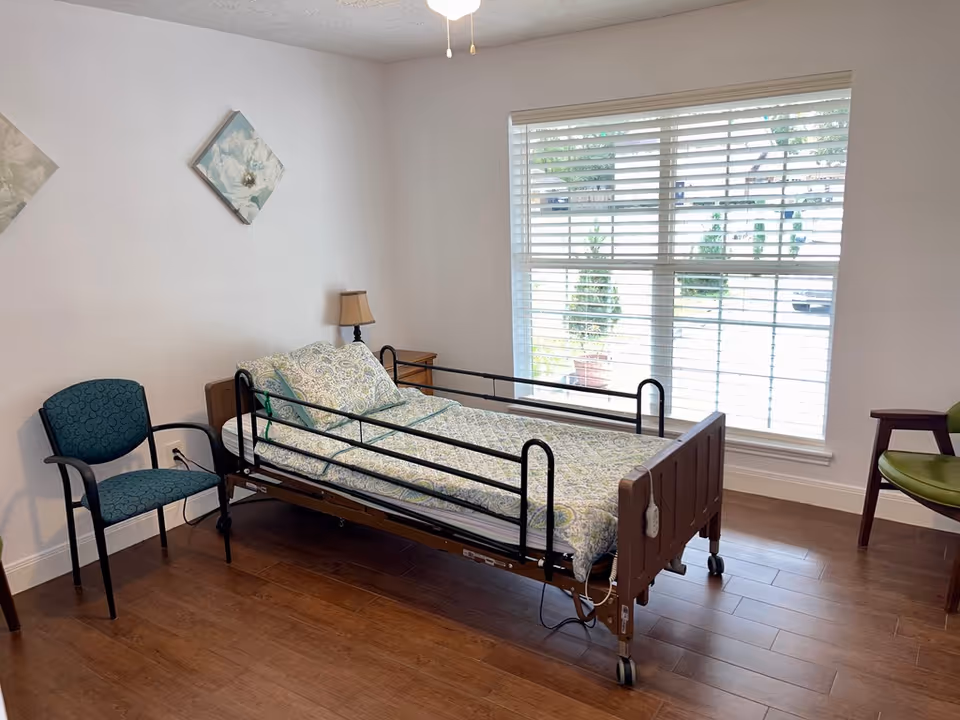 A bright assisted-living bedroom with a hospital-style bed, chairs, nightstand and a large window with blinds.