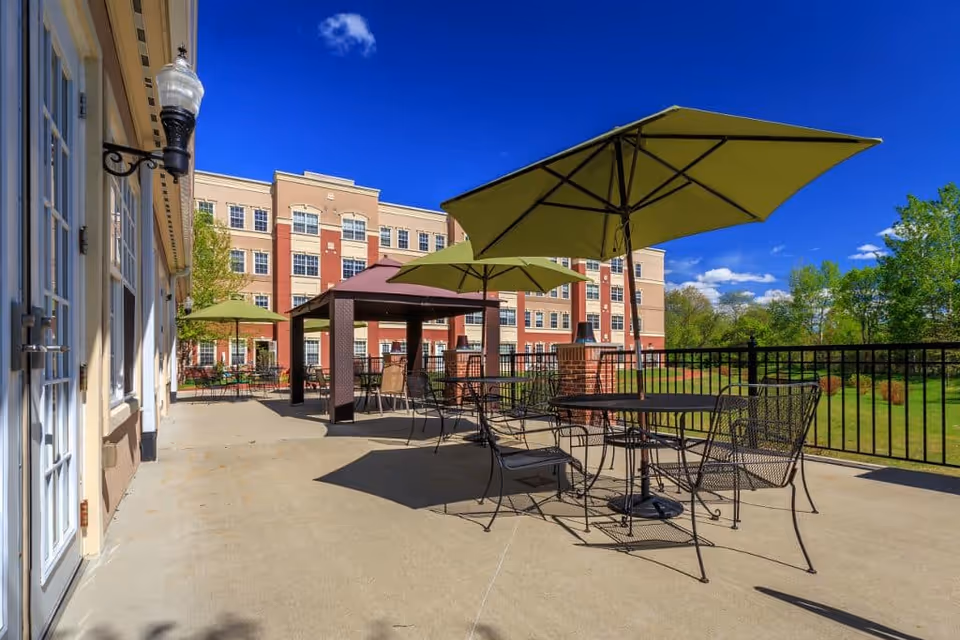Sunny outdoor patio with metal tables and chairs under green umbrellas and a gazebo in front of a multi-story senior living building.