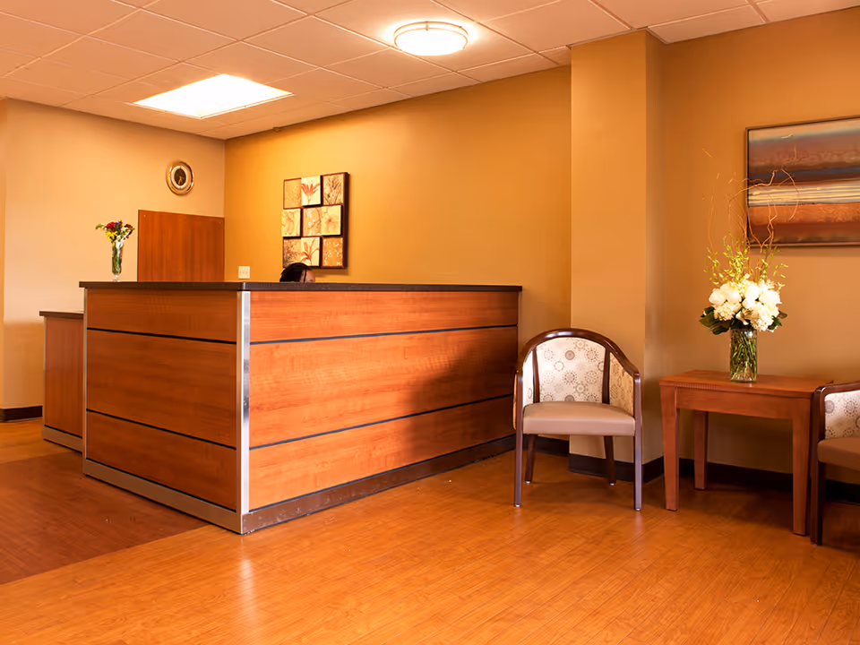 Reception area with a wooden front desk, a person partially visible behind the desk, two upholstered chairs with patterned backs, a small wooden table with a vase of white flowers, and wall art on beige walls.