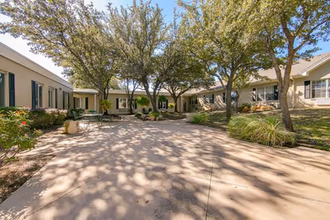 Outdoor courtyard area at Ashwood Court with paved walkways, several trees providing shade, and single-story buildings with windows and doors surrounding the space. There are some plants and bushes along the edges of the buildings.