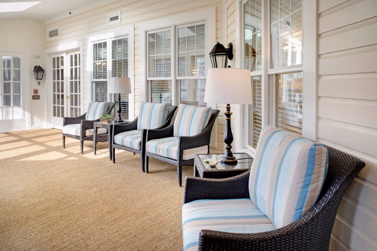 A bright and airy indoor seating area with four wicker armchairs featuring blue and white striped cushions. Each pair of chairs is separated by a small glass-top side table with a lamp. The space has large windows with white frames and white walls, allowing natural light to fill the room.
