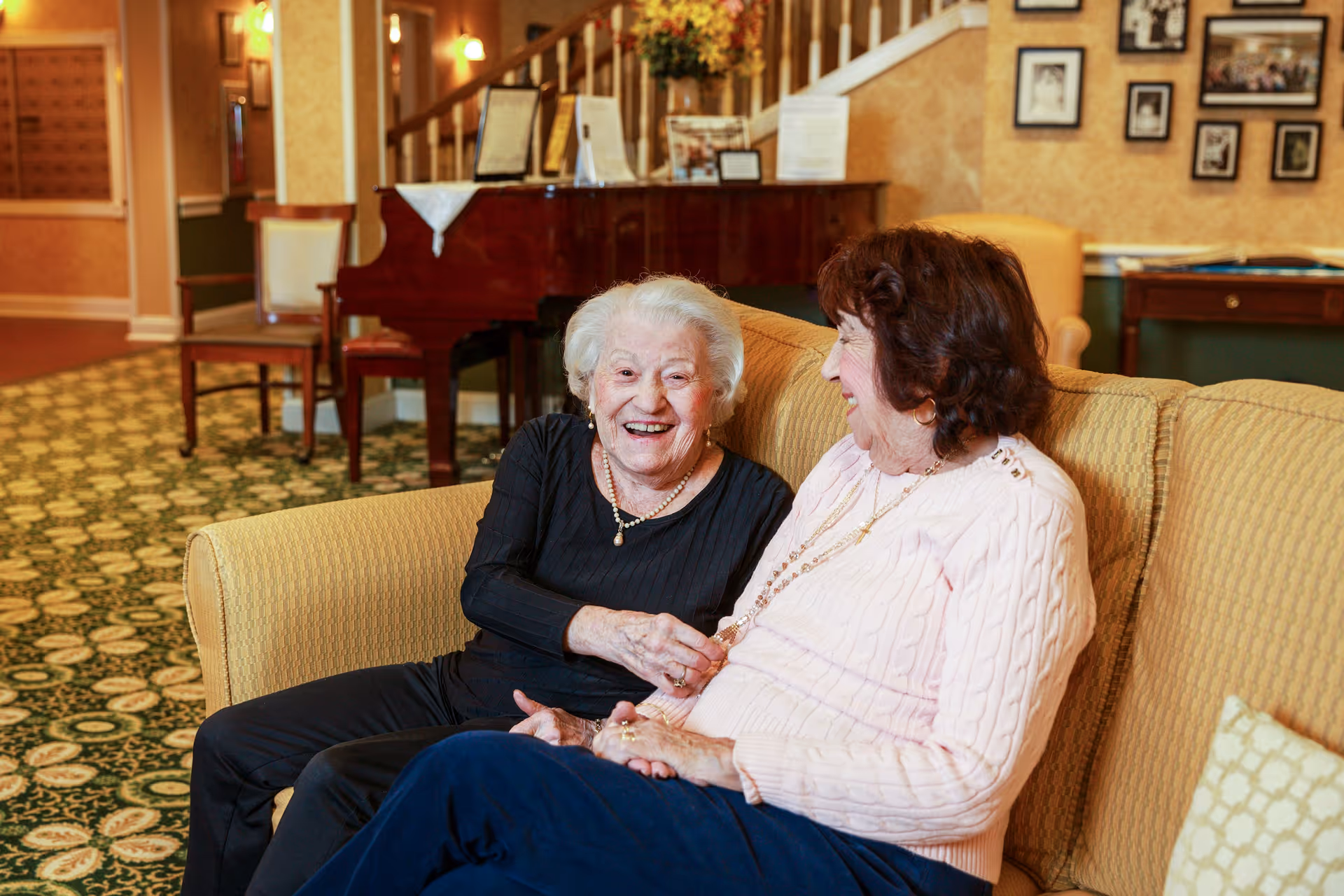 Two elderly women sitting on a yellow couch in a cozy living room, smiling and engaging in conversation. Behind them is a grand piano with a floral arrangement on top, a staircase, framed pictures on the wall, and warm lighting.