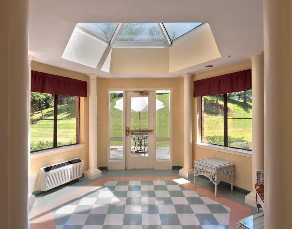 A bright interior room with large windows on both sides, cream-colored walls, and a checkered floor in light blue and white. The room features four white columns and a glass door leading outside to a patio area with a table and umbrella. A skylight above allows natural light to fill the space. There is a white wicker table and a heating/cooling unit beneath one window.