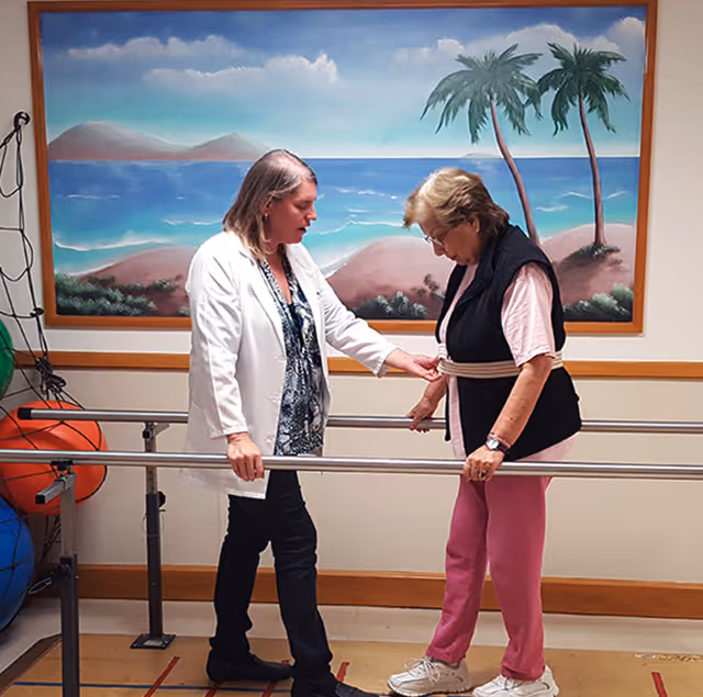 A healthcare professional assists an elderly woman with walking exercises using parallel bars in a therapy room. The elderly woman is wearing a safety belt and casual clothing. Behind them is a large painting of a beach scene with palm trees and mountains.