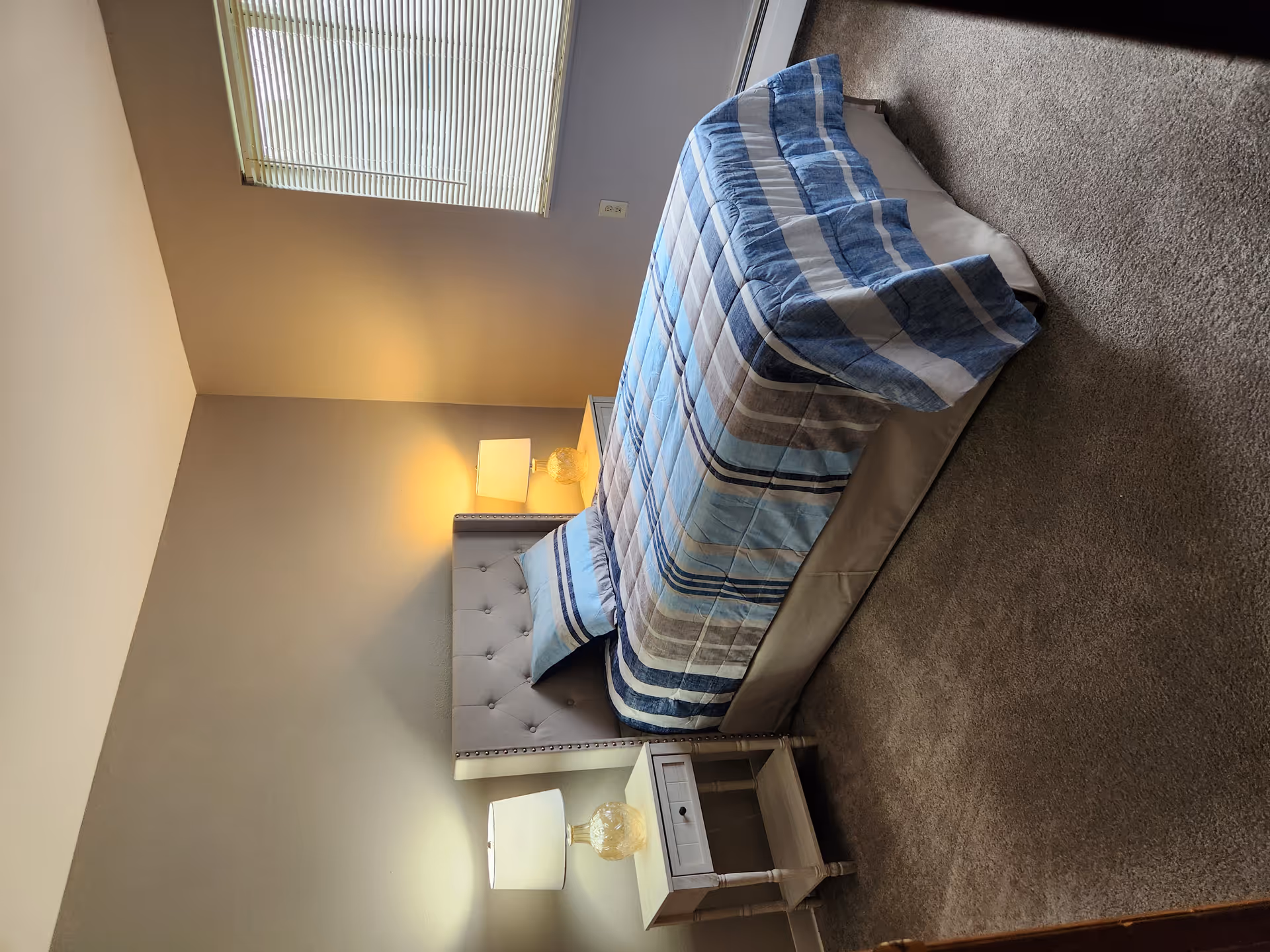 Bedroom with a bed dressed in blue striped bedding, two nightstands with lamps, a window with blinds, and carpeted floor.