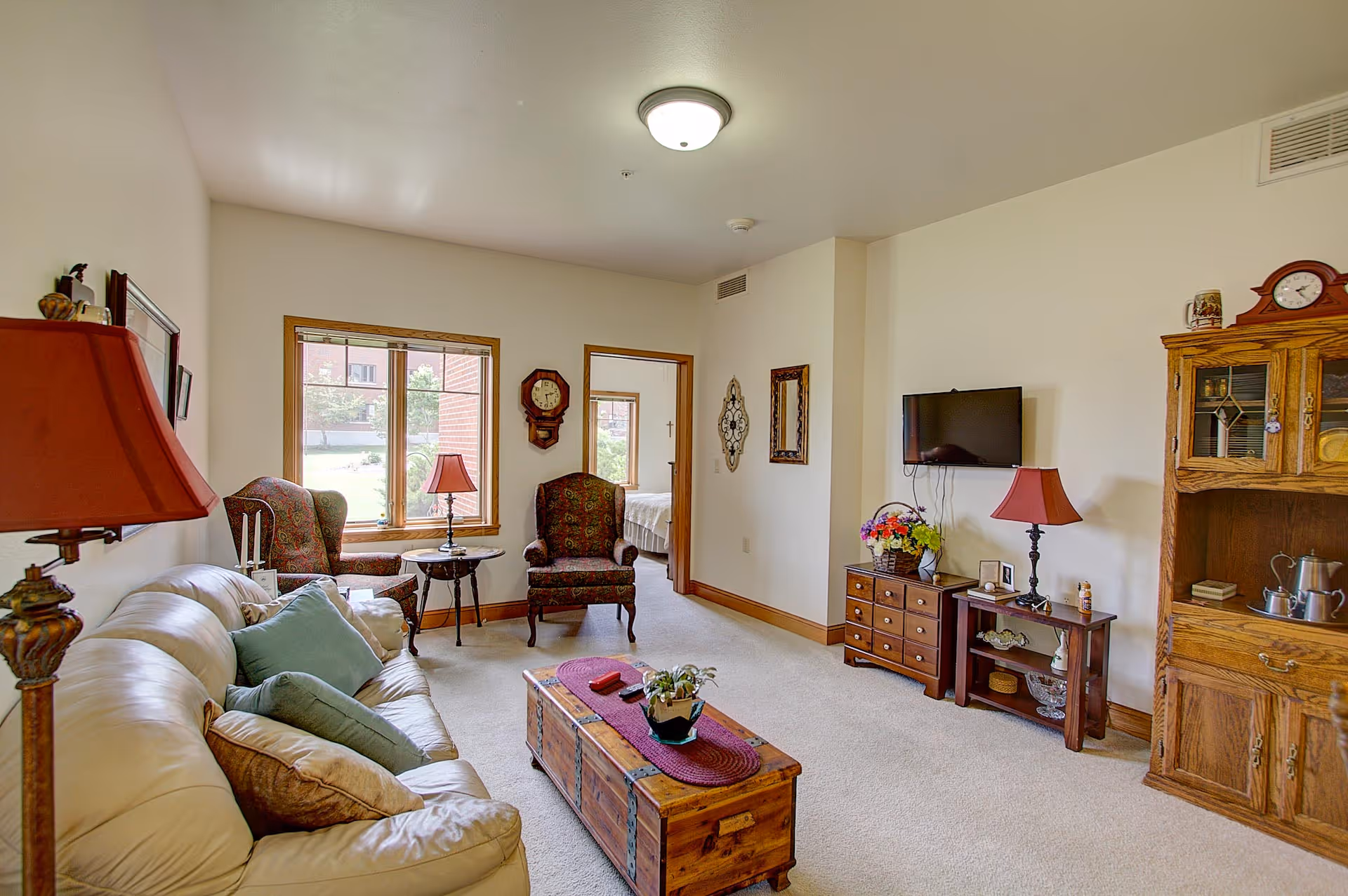 A cozy living room in a senior living community featuring a beige leather sofa with multiple cushions, two patterned armchairs near a window, a wooden coffee table with a plant and decorative runner, a wall-mounted flat-screen TV, and wooden furniture including a cabinet and side tables with lamps and decorative items.