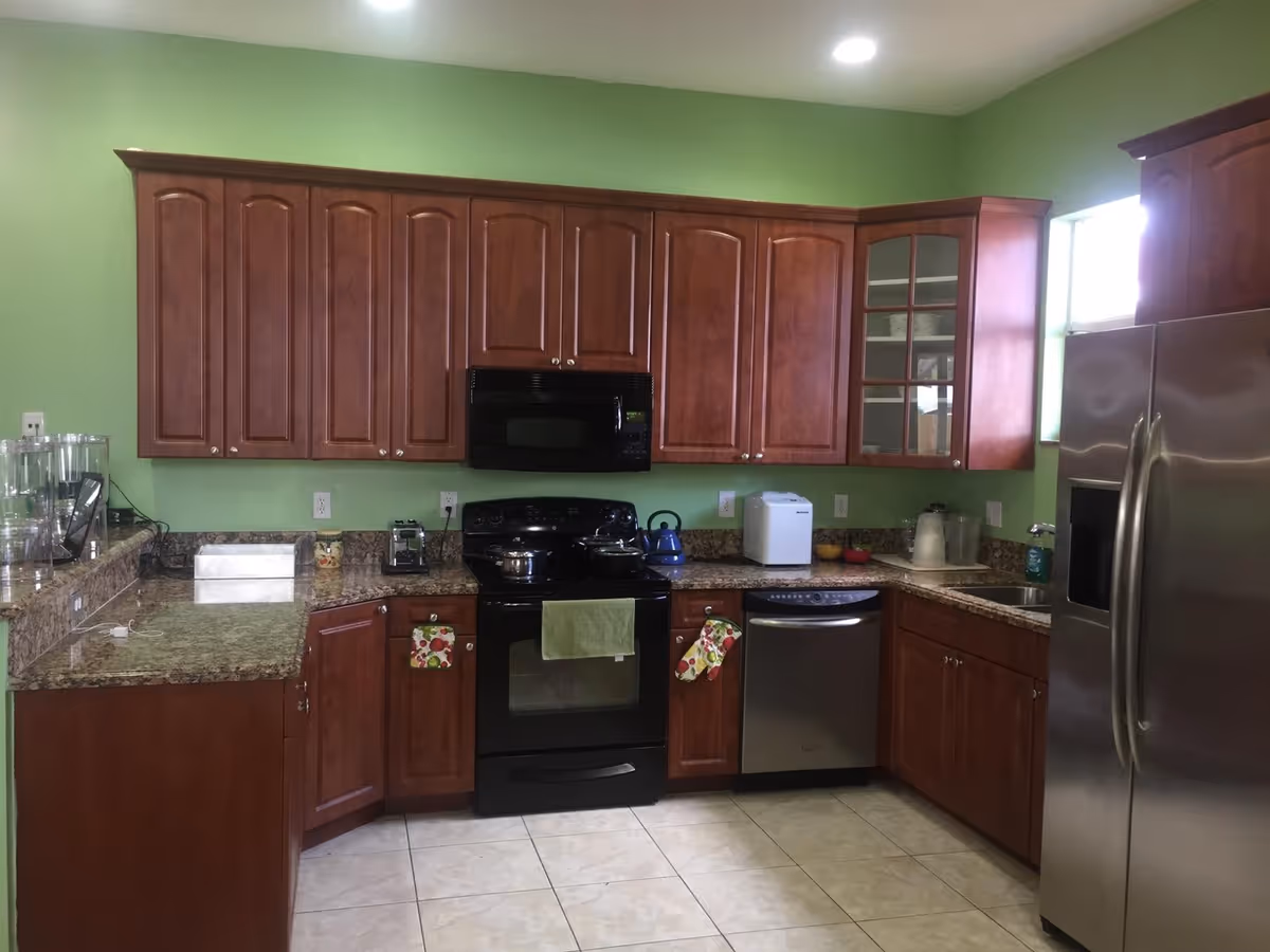 A kitchen with green walls, brown wooden cabinets, granite countertops, a black stove with a green towel hanging on the handle, a stainless steel dishwasher, and a stainless steel refrigerator. Various kitchen appliances and utensils are placed on the countertops.