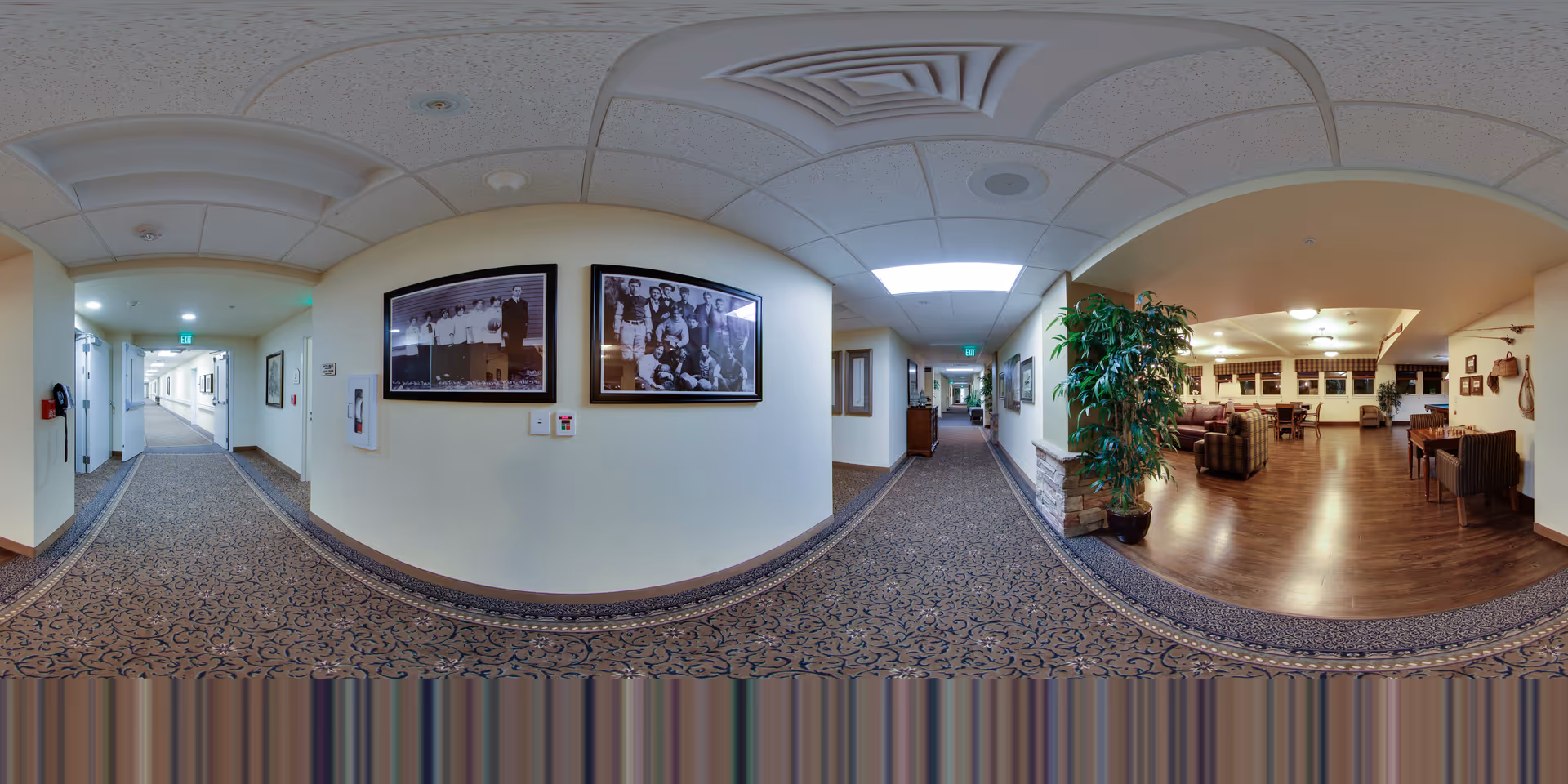 A wide panoramic view of an interior hallway in a senior living facility. The hallway has patterned carpet and cream-colored walls adorned with framed black and white photographs. To the right, there is an open living room area with wooden flooring, comfortable seating including armchairs and sofas, a large potted plant, and dining tables with chairs. The ceiling has recessed lighting and a drop ceiling design.