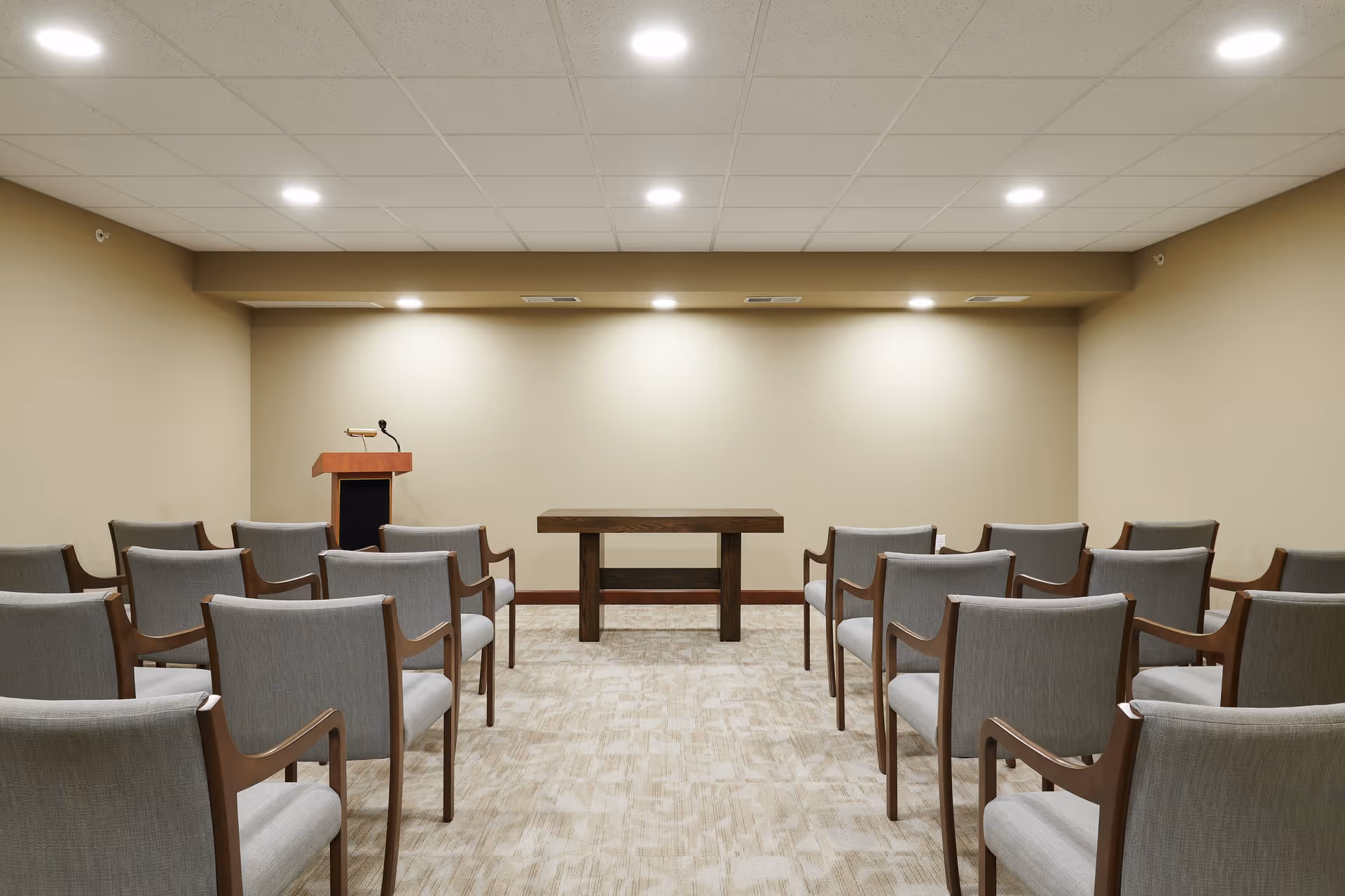 A small conference or meeting room with rows of gray upholstered chairs with wooden arms facing a wooden podium with a microphone and a small wooden table against a beige wall under bright ceiling lights.