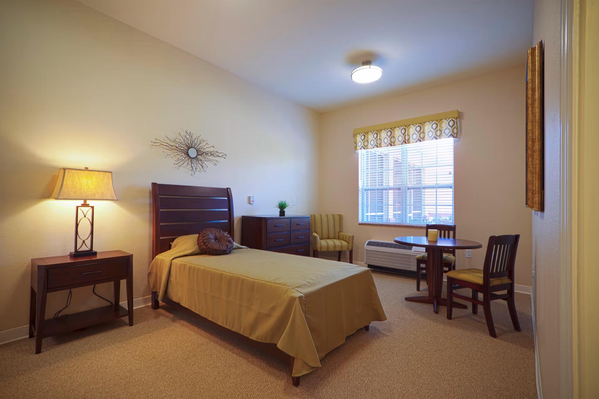 A neatly arranged bedroom in an assisted living facility featuring a single bed with a brown bedspread and round pillow, a wooden nightstand with a lamp, a wooden dresser with a small plant on top, a striped armchair, a round wooden table with two chairs, and a window with blinds and a patterned valance.