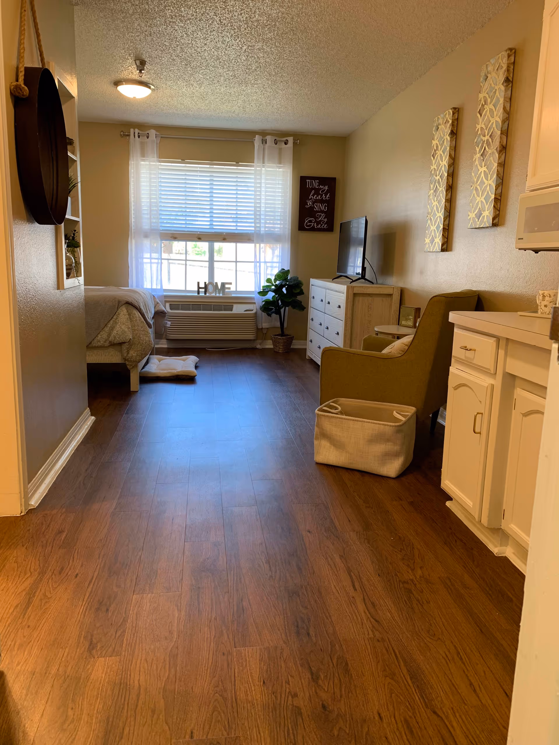 Interior view of a cozy senior living apartment at Bradfield Terrace featuring a wooden floor, a bed partially visible on the left, a window with white curtains and blinds, a small air conditioning unit below the window with decorative letters spelling HOME on it, a green armchair with a beige basket beside it, a white dresser with a TV on top, and wall decorations including two patterned panels and a framed quote.
