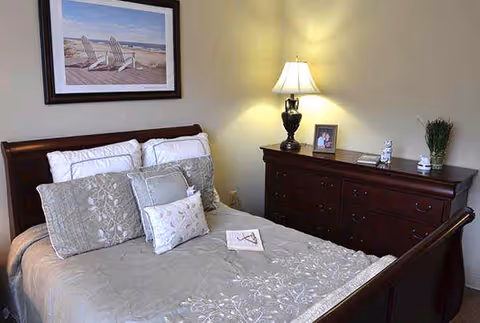 Neatly made bed with decorative pillows, a wooden dresser with a lamp and framed photo, and beach artwork on the wall in a bedroom.