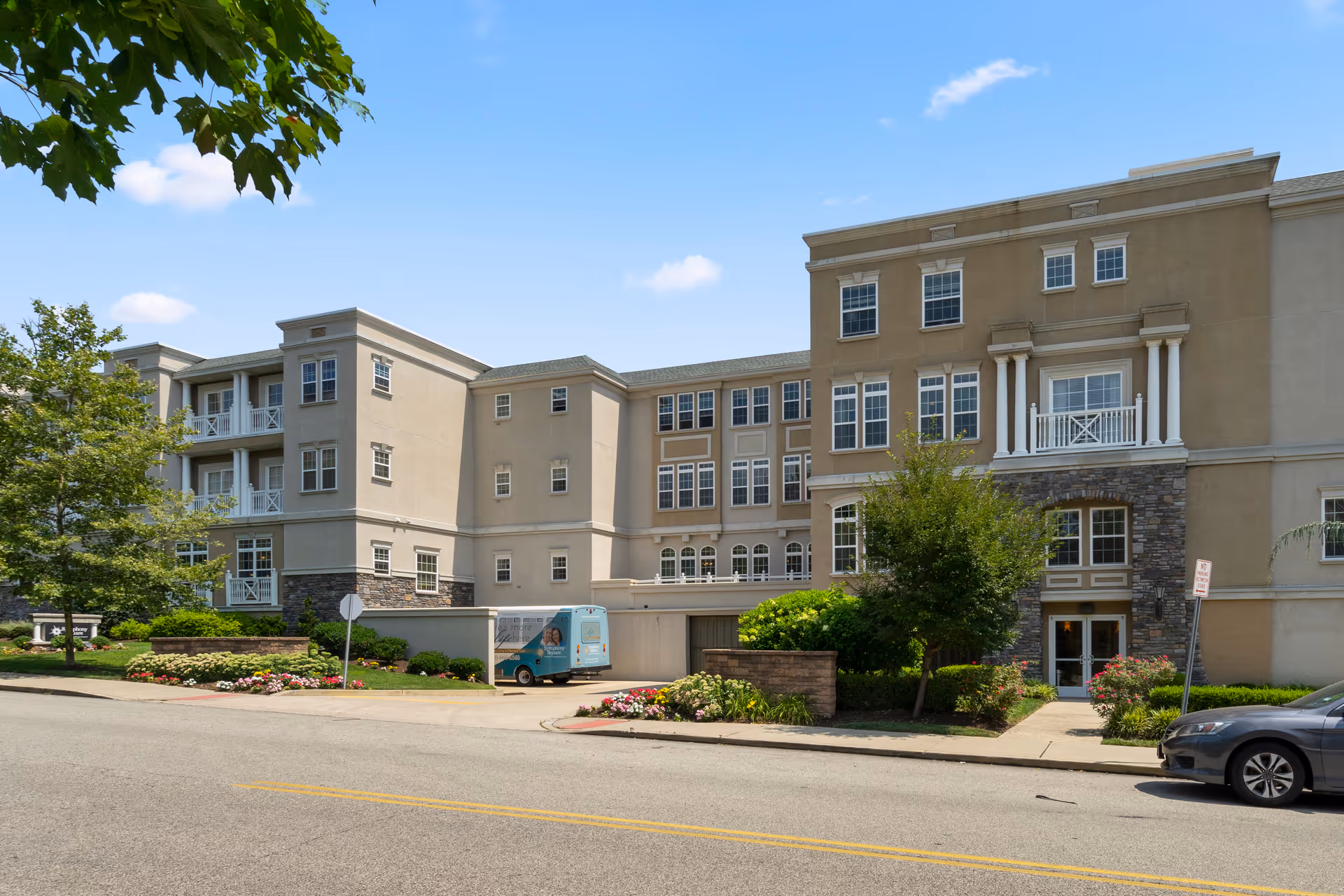 Exterior view of a multi-story senior living facility building with beige walls, multiple windows, balconies, and landscaped greenery including trees and flower beds. A blue van is parked near the entrance, and a car is parked on the street in front of the building under a clear blue sky.