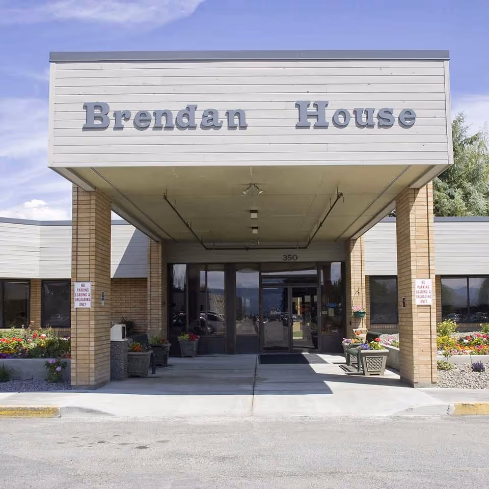 Front entrance of Brendan House, a single-story building with a covered drop-off area supported by brick pillars. The building name 'Brendan House' is prominently displayed above the entrance. There are flower beds and planters with colorful flowers on either side of the entrance walkway.