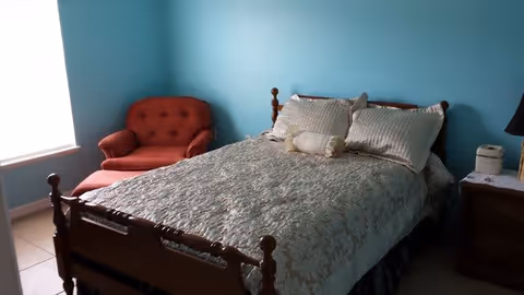 A bedroom with a wooden bed frame, a bed covered with a patterned bedspread and pillows, a red upholstered armchair near a window with white blinds, and a wooden nightstand with a lamp and a small white appliance.