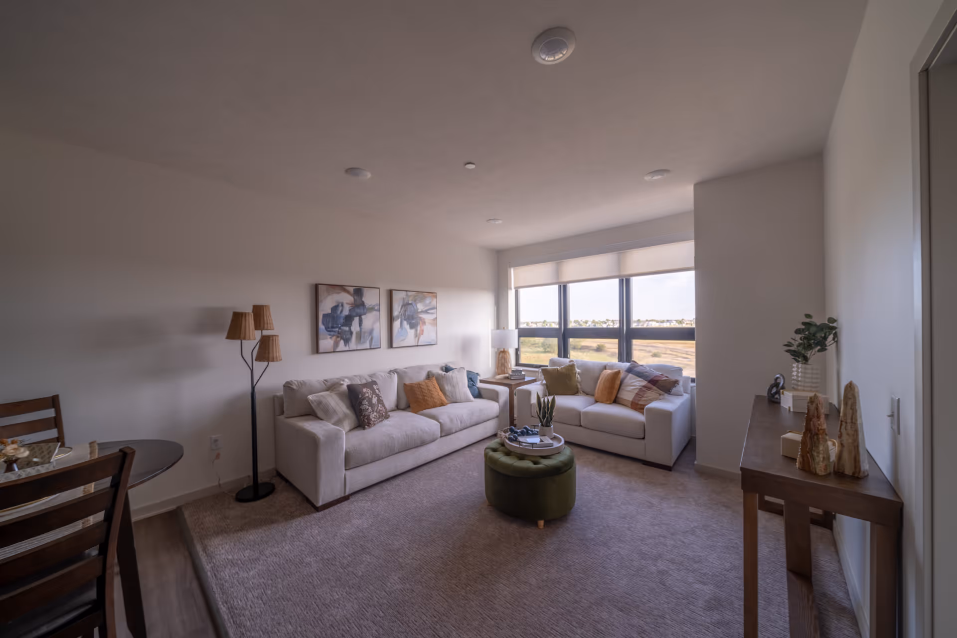 A cozy living room with two white sofas adorned with various throw pillows, a round green ottoman in the center, a floor lamp with three wicker lampshades, a wooden side table with decorative items, and large windows letting in natural light with a view of an open field outside.