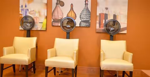 Interior view of a salon area with three vintage-style hair dryer chairs with beige upholstery against an orange wall. Above the chairs are three framed pictures featuring decorative glass bottles.