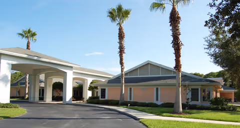 Exterior view of a single-story building with a covered entrance driveway, surrounded by palm trees and well-maintained landscaping under a clear blue sky.