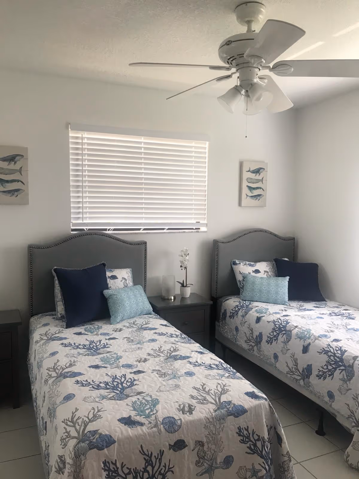 A bedroom with two twin beds featuring matching headboards and bedding with a sea life pattern. Each bed has decorative pillows in shades of blue. Between the beds is a nightstand with a small potted plant and a candle. Above the beds are two framed pictures of fish. A ceiling fan with lights is mounted on the ceiling, and a window with white blinds is on the wall behind the beds.