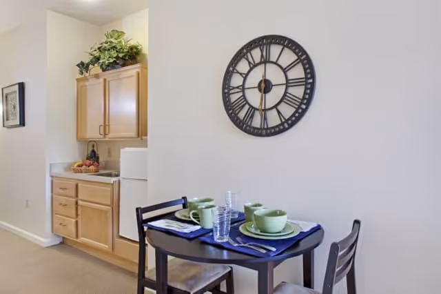 Small dining area with a dark wooden table set for two with green dishes, cups, and clear glasses on blue placemats. Behind the table is a wall with a large black clock. To the left is a kitchenette with light wood cabinets, a white refrigerator, and a countertop with a bowl of fruit and a small statue. A framed picture hangs on the wall near the kitchenette.