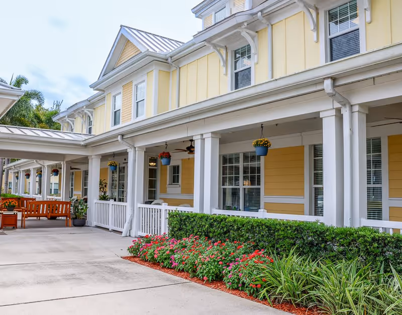 Exterior view of a senior living facility with yellow siding and white trim. The building has a covered porch with white railings and hanging flower pots. There are benches and potted plants along the walkway, and a neatly maintained garden with green shrubs and colorful flowers in front.