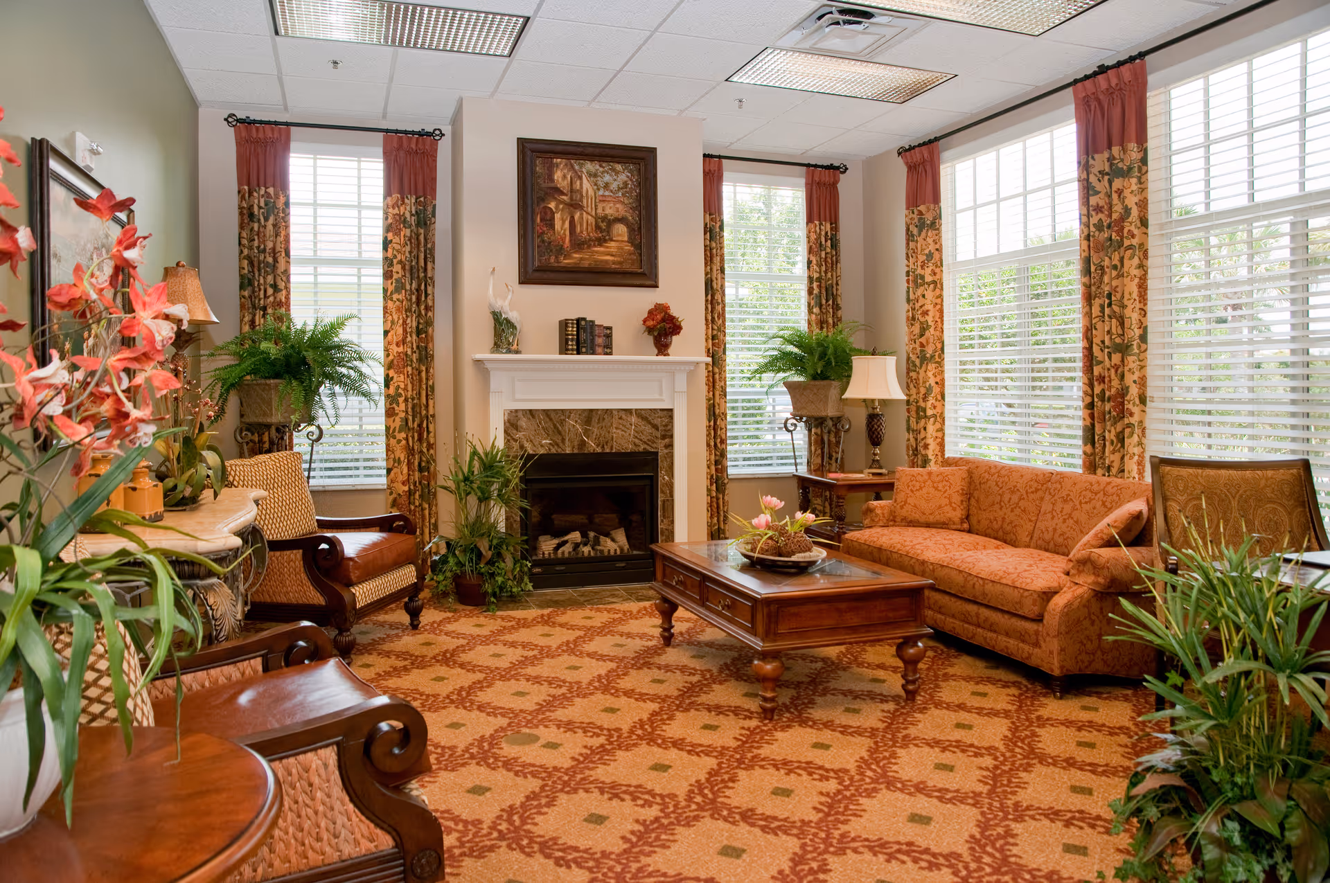A cozy living room with a patterned carpet, an orange sofa, two armchairs, a wooden coffee table with a floral centerpiece, and a fireplace with decorative items on the mantel. The room has large windows with floral curtains and several green plants adding to the warm ambiance.
