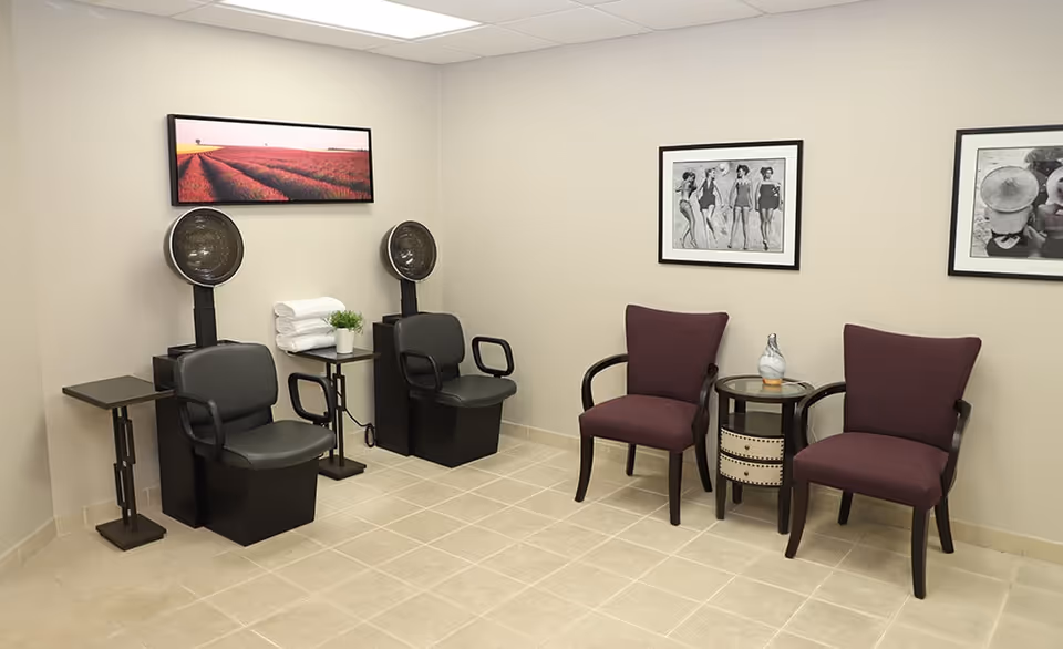 Small salon/waiting area with two hooded hair dryers, two black salon chairs, two purple guest chairs and framed artwork on the walls.