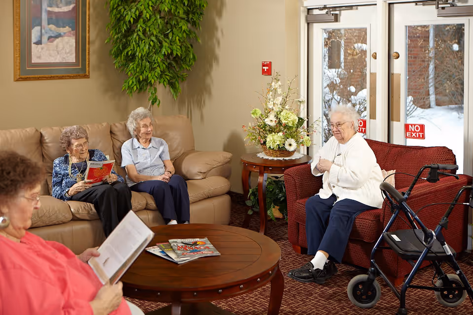 A cozy living room area in St Joseph Residence with four elderly women. Three women are seated on a beige couch, two of them reading magazines, and one woman is sitting on a red armchair near a walker. There is a round wooden coffee table with magazines on it, a side table with a floral arrangement, and a large plant against the wall. Glass doors with 'No Exit' signs show a snowy outdoor scene.