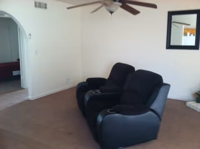Two black reclining theater seats sit in a sparsely furnished living room with a ceiling fan and arched doorway.
