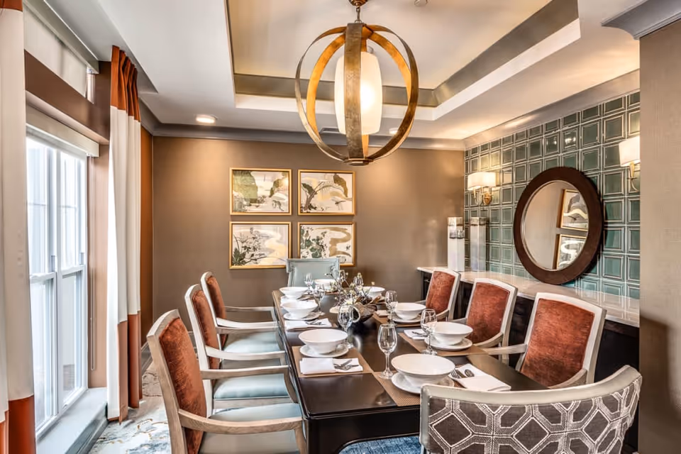 A well-lit dining room with a long dark wooden table set for eight people. The chairs have a mix of rust-colored and patterned upholstery. The room features a large window with white and rust-colored curtains, a modern circular chandelier, four framed artworks on one wall, and a round mirror on a green tiled accent wall with two wall sconces.