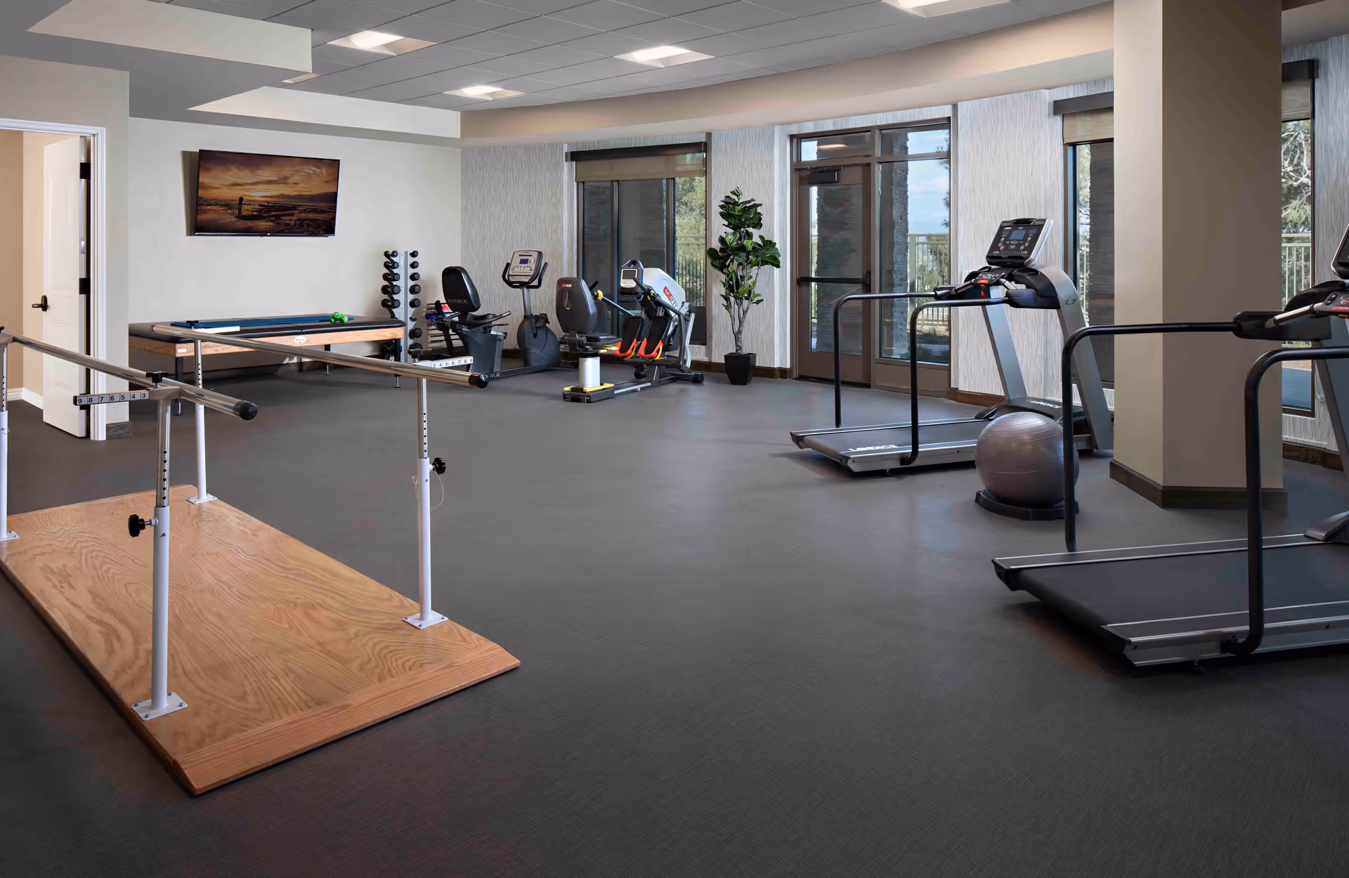 A senior living facility fitness room with exercise equipment including treadmills, stationary bikes, a balance ball, and parallel bars. The room has large windows and a door leading outside, with a wall-mounted TV and a small rack of weights.