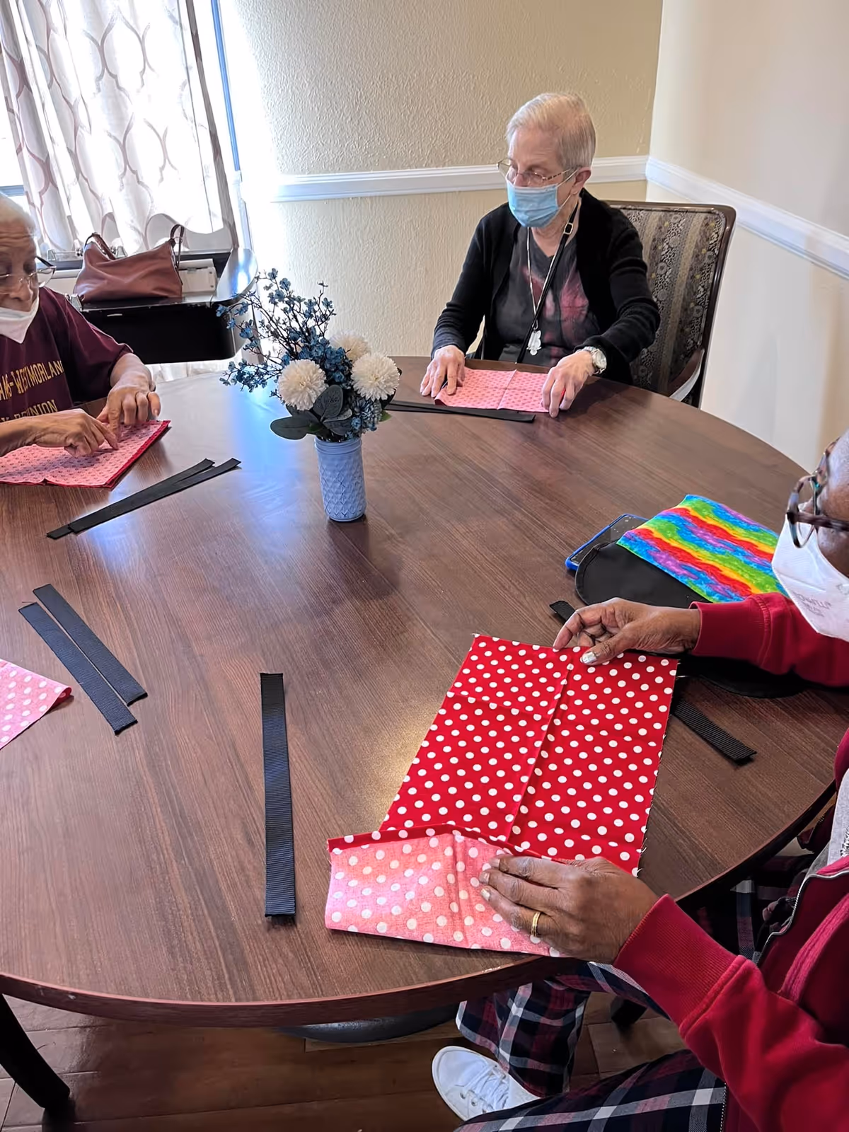 Three masked residents sit around a round table in a common room folding colorful polka-dot fabric with a small floral centerpiece.