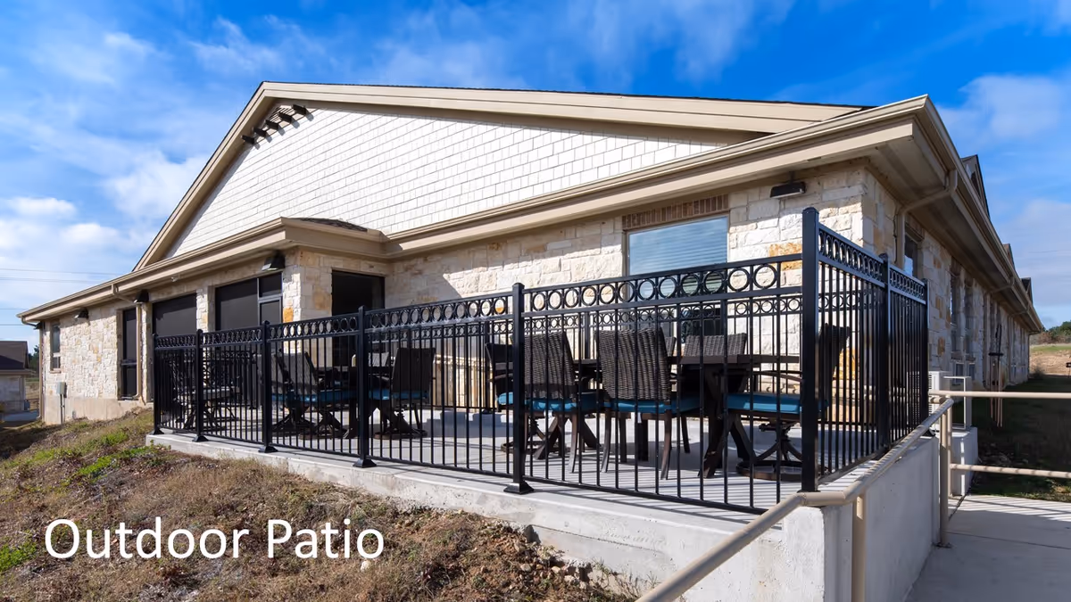 Outdoor patio area at New Haven Assisted Living and Memory Care of Bastrop featuring a stone building with a fenced patio containing multiple tables and chairs under a blue sky.