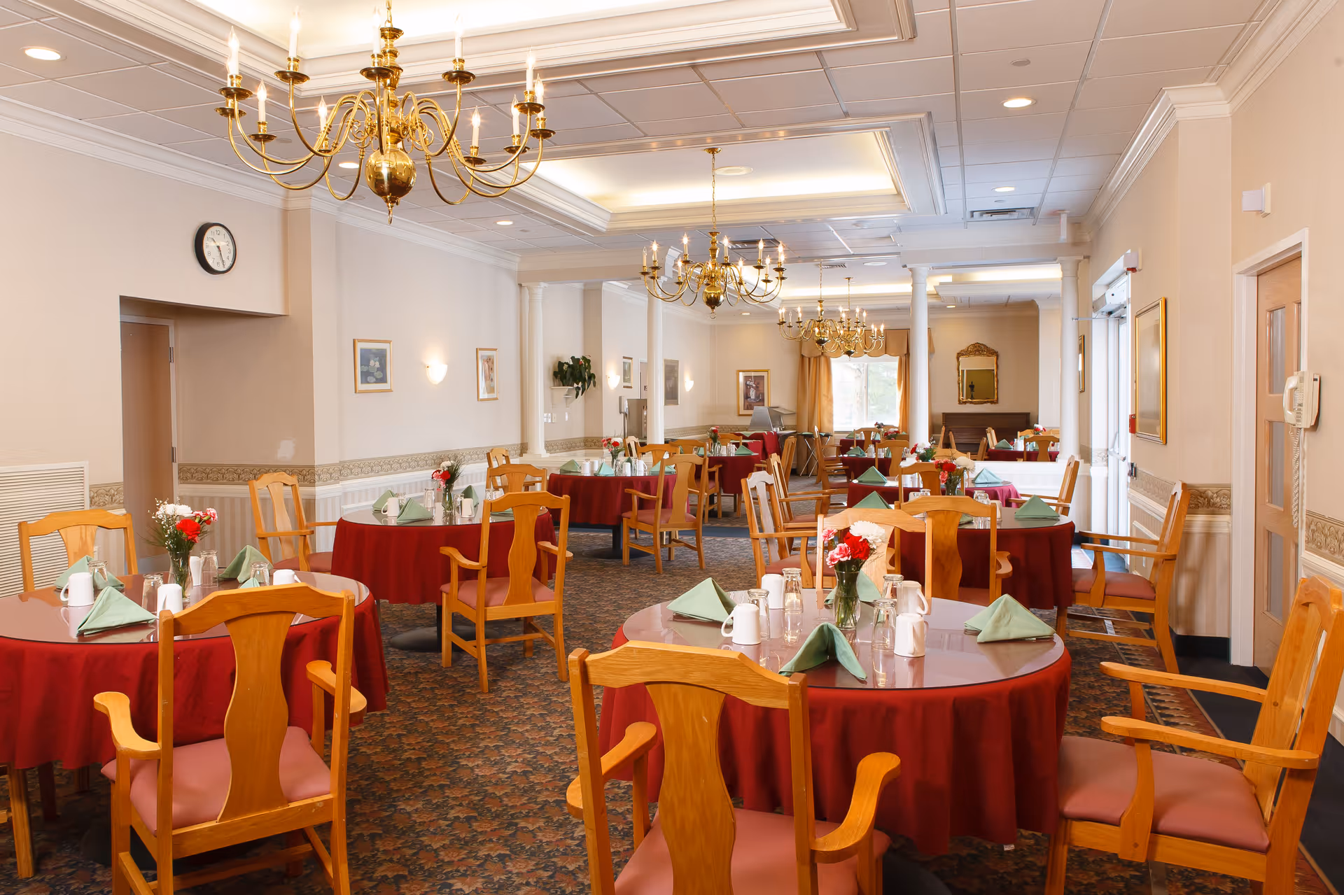 A spacious dining room with round tables covered in red tablecloths, each set with green folded napkins, white mugs, and small flower vases. Wooden chairs with armrests surround the tables. The room features ornate chandeliers hanging from a white coffered ceiling, framed pictures on the walls, and a carpeted floor with a patterned design. There are columns and large windows with curtains in the background.