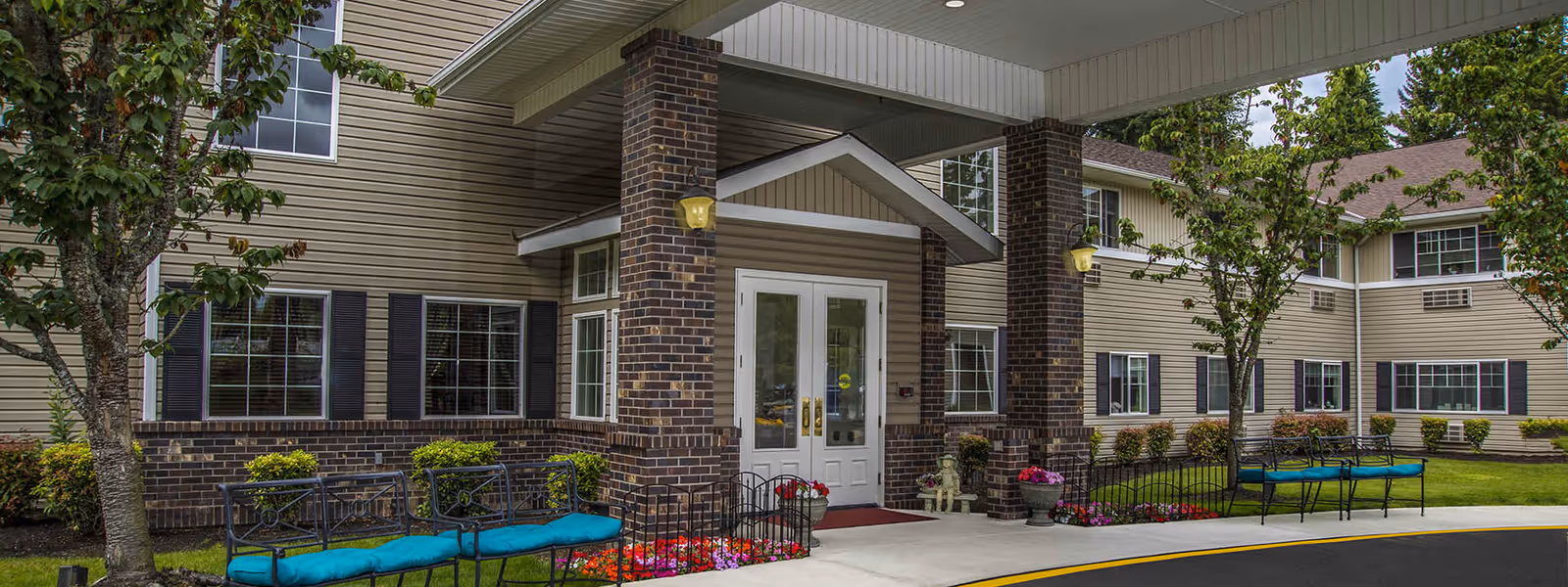 Entrance of Normandy Park Senior Living facility showing a covered porch with brick pillars, white double doors, outdoor benches with blue cushions, trees, and landscaped flower beds.