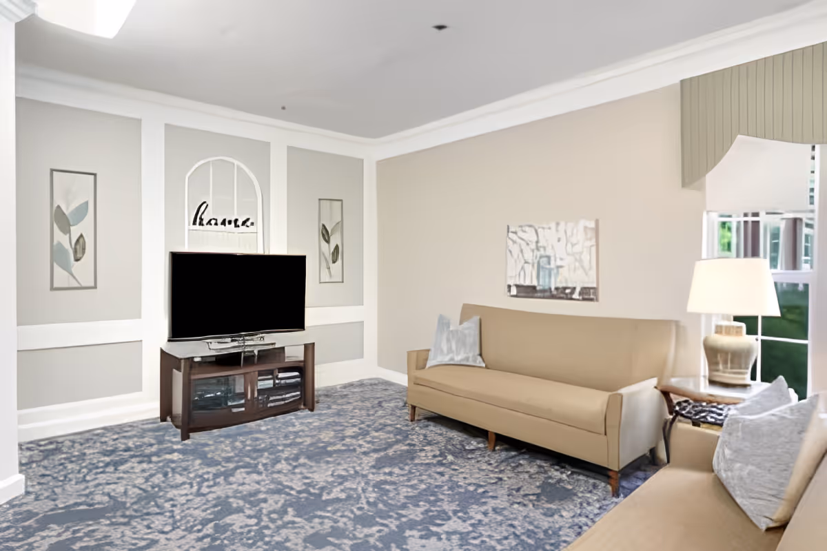 A living room with two beige sofas, a wooden TV stand with a flat-screen television, decorative wall art, a table lamp on a side table, and a large window with a valance. The room has a patterned blue carpet and light-colored walls.